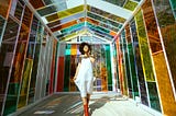 A woman stands smiling in a multicolored, stained glass atrium wearing a white dress and red boots.