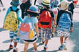 Young school children crossing a street from the back, multi-color backpacks and wide brimmed hats can be seen.