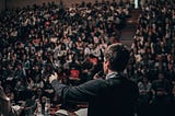 A man in a black suit has his back turned as he addresses a crowd inside a theater