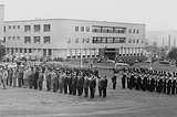 Black-and-white photograph of officials standing in lines outside buildings.