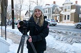 Lauren Weseluck holds a camera on a tripod on the sidewalk with the street and older-style houses in the background.