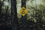 Two trees close up in a forest with sun shining between trees. A yellow diamond shaped sign with Stay On Trail is fixed to one of the trees