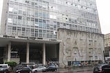 A imagem mostra a Avenida Paulista à frente do prédio da Gazeta, com alguns carros passando. Ao fundo é possível ver o prédio. No seu lado esquerdo temos um escadão e ao lado direito uma gigantesca parede escrito Gazeta três vezes.