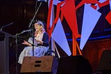 image of a woman in a wheelchair on stage reading, she is wearing a tutu, glasses and has long pink curly hair