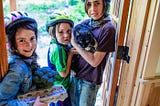 Three smiling children in bicycle helmets, standing in a doorway, one holding a cat and another holding a carton of eggs and broccoli.
