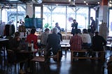 A speaker is standing and holding a microphone, whilst talking to another who is seated. They are in front of a large window that has a small round sticker that reads “Ethical Dilemma Cafe”. Clusters of audience members are seated on chairs and wooden benches in front of them.