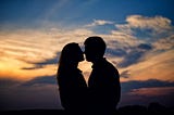 Woman and man face to face silhouetted against sky at sunset