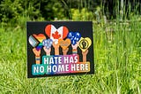 A sign saying, “Hate has no home here” and various flags, in the middle of long grass.