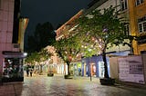 A downtown street at night with very few people and illuminated only by lights from stores and streetlamps.