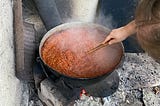 A hand stirs a large pot of sauce over a fire.