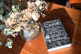 The book Caste: The Origins of Our Discontents by Isabel Wilkerson next to a vase with wilted white roses on a wooden table