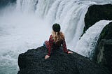 A woman sitting on edge of cliff on waterfall.