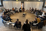 A group of people sit in white chairs in a circle around a room. In the center of the room are candles. The room is lowly lit with grey walls flanked bywhite panels.