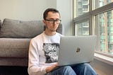 A man in his mid 20s wearing a white sweather and blue jeans. He’s sitting on the floor against a couch. He’s resting a MacBook against his lap. He has black hair and glassses. On the right-hand side of the frame is a window.