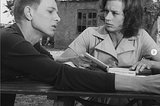 Screenshot from Ingmar Bergman’s Through A Glass Darkly. A young man and woman sit at a picnic table, the woman holds a book in her hands. Subtitle reads: You in your cage, I in mine.