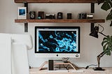 An Apple desktop on a table with a lamp, shelves, and a few Monstera leaves.