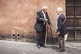 An Elderly Man With Walking Stick Speaking To An Elderly Woman Outside A Building In Europe.