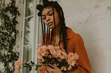 A woman with Box braids leans up against the wall, holding a bouquet of flowers.