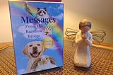 A box of oracle cards leans against a lamp on an end table with a figurine next to it of a blond haired girl with wings on her knees praying. The oracle card shows a blue sky with white cloud and a rainbow with pictures of a bird, cat, hamster, dog and rabbit. The box says: Messages From the Rainbow Bridge. Oracle Cards.