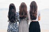 Three slim brunette women stand close with their backs to the camera, looking towards a misty landscape. Two wear long flowing dresses, the other a long black skirt and tight white top.