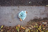 A dirty blue surgical mask lays on the ground, crumpled up next to a pile of leaves.