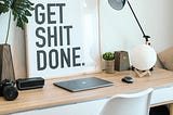 photo of a desk with a poster leaning against the wall that says get shit done