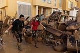 Two women wearing boots full of mud and walking on a pavement all brown due to the mud are carrying one bucket (one from each handle, with the bucket in between them). Behind them, very close, is a big pile of mud-covered debris. In the background, the houses of people in light sand-colour rendered walls.