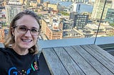 Lisie wearing a Google accessibility shirt, with a beautiful view of Manhattan behind her. The shirt says Google, but with the ‘o’s written in braille, the second ‘g’ represented in sign language, and the ‘e’ shown with a person sitting on it as if the letter were a wheelchair