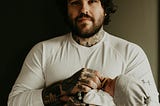 An adult man with a beard and tattoos holds a pacifier to an infant’s mouth.