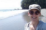 Photo of author, Gill McCulloch standing on a sandy beach in Sooke, BC, Canada. The sun is shining on waves with trees in the background.