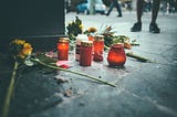 Candle and flowers on the ground in a public street as people walk by and one person stands by them
