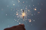 A hand holding a stick giving off little sparks (a firework sparkler) at night