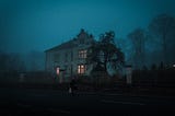 The image shows a large house at dusk. The trees behind the property are shrouded in fog. Some of the windows are lit, others are dark. A lone figure is crossing the road to approach the house.