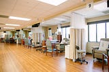 Five consecutive beige hospital chairs are separated by cream curtains pulled back with one extra seat for each station and a rolling charting cart.