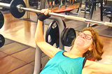 woman using the incline chest press grimaces as she lifts the bar with five pound weights on either end