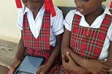 Two girls in school uniform sit outside on a wooden bench. One holds a tablet and the other has her arms crossed. Both are looking at the tablet.