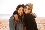 Two young women, best friends, embracing on the beach, one with her arm around the shoulder of the other, the two of them, heads touching, smiling, looking out at the camera.