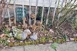 Weathered stone goose garden statue lying on its side in a leaf-covered flower bed bordered by old bricks