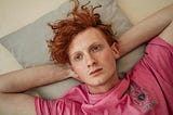 Young man lying on his back looking up, red hair pink shirt