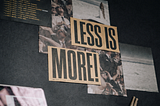 Image of a wall with images, text stuck to it with two big paper cards that read “Less is more”.