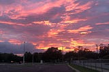 Photo features a sunset with orange and purple colored sky and many clouds. Beneath sky is a dark parking lot.
