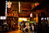 A night shot of a very busy corner in central Hong Kong. Neon signs shine bright from various buildings. A white bus with green top is turning around the corner.