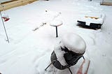 A snowy backyard scene shows garden features covered in a thick blanket of snow. A bird feeder stands in the foreground, also topped with snow, emphasizing the quiet stillness of the winter setting. Footprints dot the snow, adding a hint of motion amidst the serene landscape.