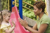 White child with long blond hair/pigtails playing paddycake with black women with short hair and green shirt on playground