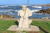 White statue of Saint James holding an iron staff. Statue is sitting on green grass. Ocean waves crash over rocks in the background.