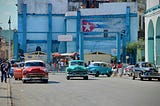 Vintage cars in bright colors, including red, green, and blue, parked on a street in Havana, Cuba. In the background, a faded blue building with a mural of the Cuban flag, and people walking along the sidewalk.