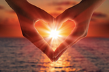 Two hands making heart shape with ocean in background and sunset shining through the centre