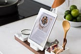 A recipe open on a tablet device supported on a tablet stand on the kitchen counter alongside a spoon and a bowl of limes