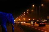 An ultramarine elephant watches cars go by on a busy highway at night, bathed in orange light