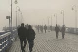 Silhouettes of people walking along a wooden pier in mist with street lamps on each side.